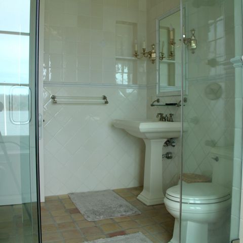 Glass shower, pedestal sink and toilet in a white tile bath with a water view
